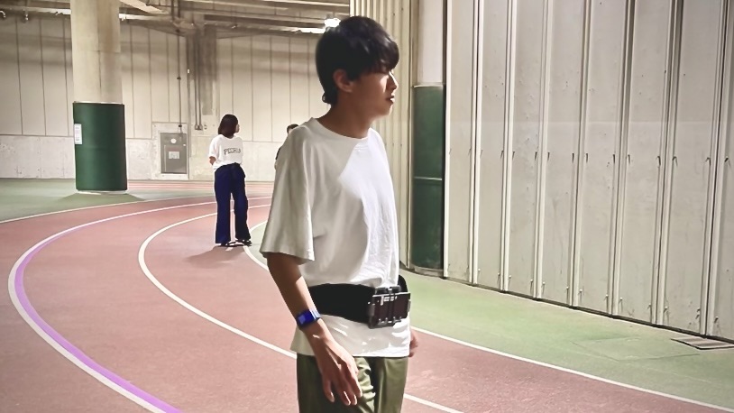 画像には、屋内のランニングトラックが映っています。前景には、白いTシャツと緑のパンツを着た男性が立っており、腰にはスマートフォンが装着されたベルトを巻いています。男性は腕に青いバンドも装着しています。背景には、別の人がスマートフォンを操作しながら立っており、さらに奥には柱や設備が見えます。 ※このALTの作成には、GCStudio（X：@GCStudio2024）によって提供されるGPTs（VisionAssist ALT）を利用しています。本ALTに関するご質問や問題がある場合は、当スタジオまでお問い合わせください。